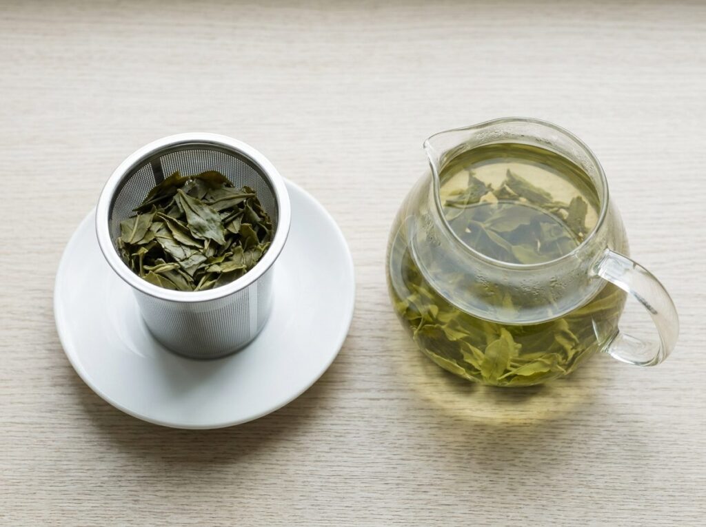 Filtre en inox rempli de feuilles de thé vert déployées, posé à côté d'une théière en verre sur un plan de travail clair