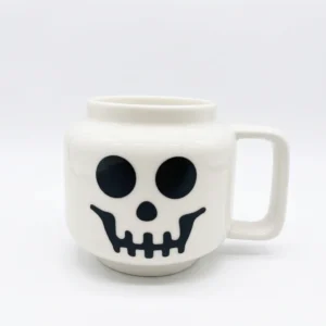 509637c4-87f4-4322-b59d-f799e2b354e2.webp Mug en céramique blanche représentant un crâne souriant avec une anse rectangulaire classique